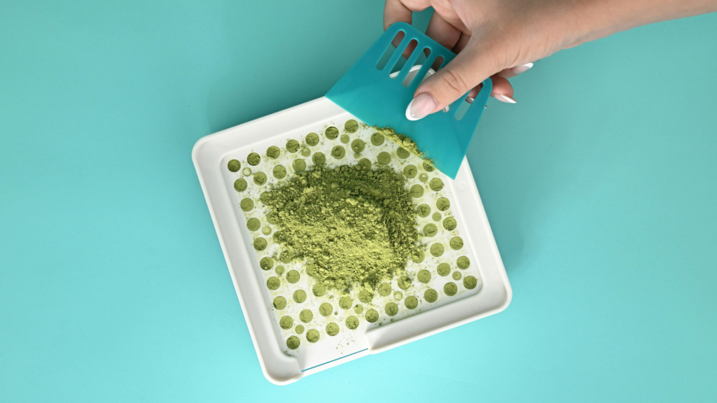 Hand using a scraper to spread powder evenly across a capsule filling tray