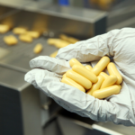 Gloved hand holding filled capsules beside a capsule filling machine tray.