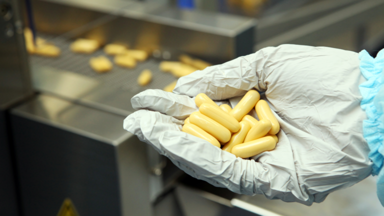 Gloved hand holding filled capsules beside a capsule filling machine tray.