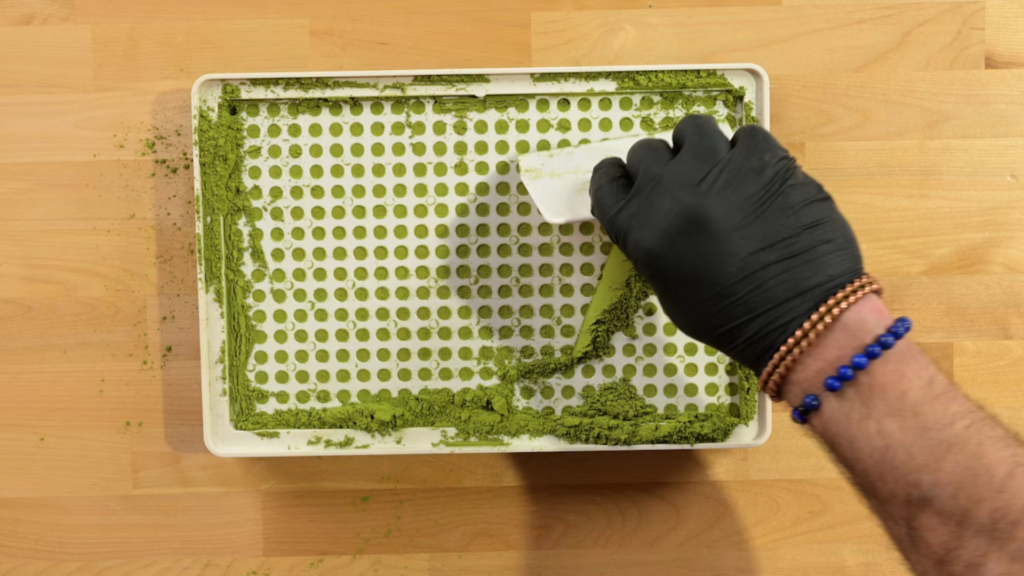 Gloved hand scraping excess powder from a capsule filling machine tray during cleaning