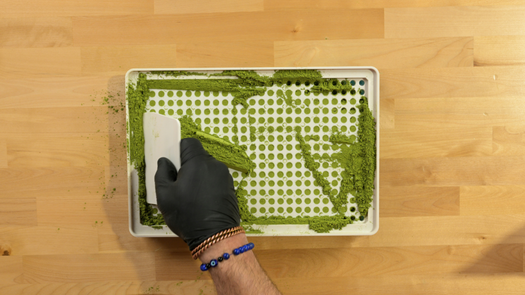 Gloved hand using a scraper to tamp green powder into a capsule filling tray