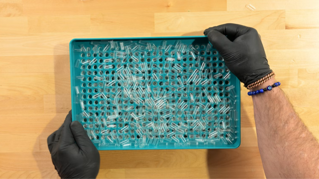 Gloved hands loading a capsule filling tray for a liquid capsule filling machine batch.